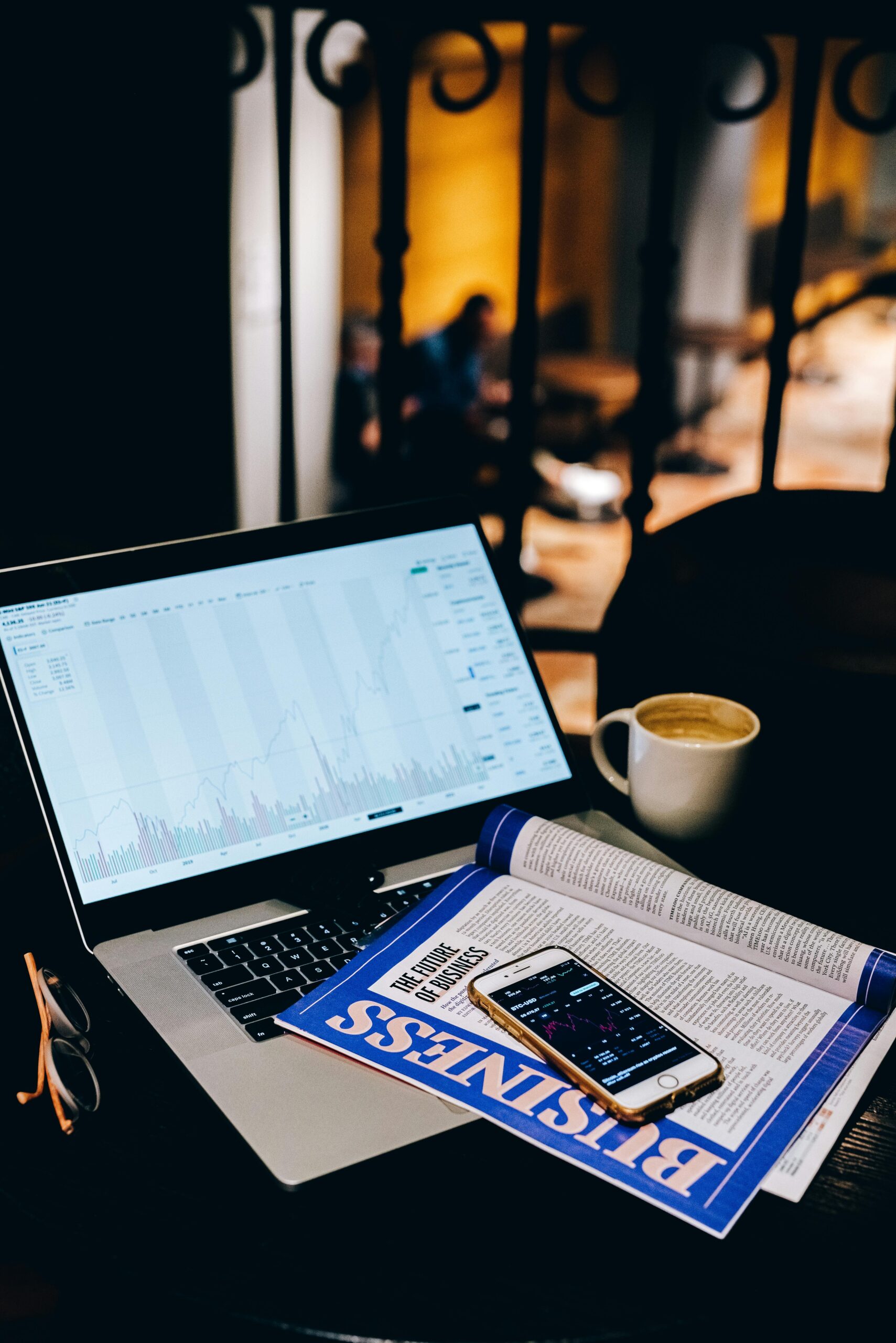 A laptop showing financial charts beside a business magazine and coffee cup in a cafe setting.